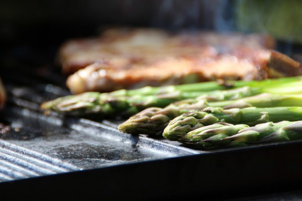 Stangen grünen Spargels auf einer Grillplatte, im Hintergrund ein Steak