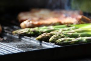 Stangen grünen Spargels auf einer Grillplatte, im Hintergrund ein Steak