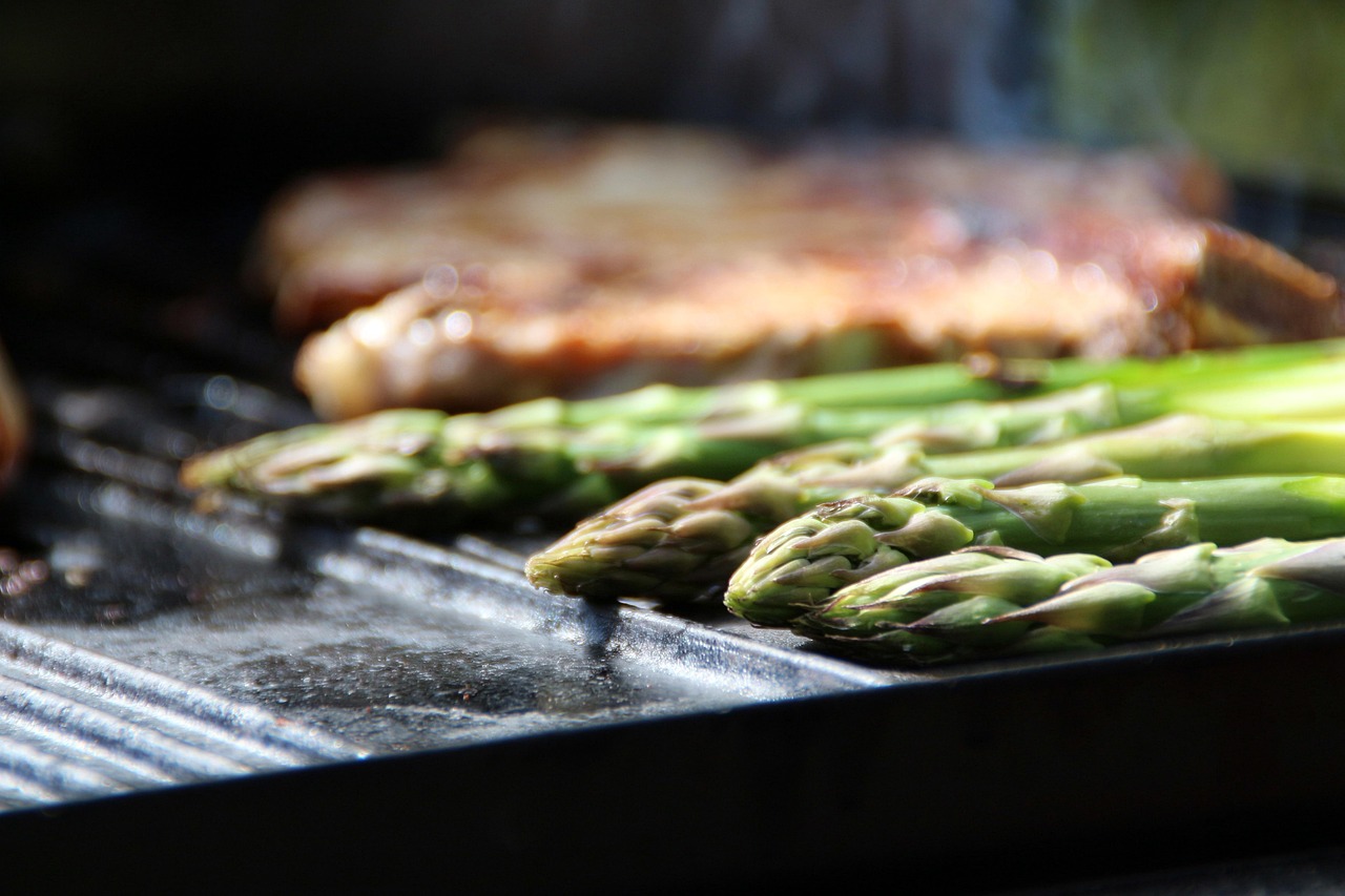 Stangen grünen Spargels auf einer Grillplatte, im Hintergrund ein Steak