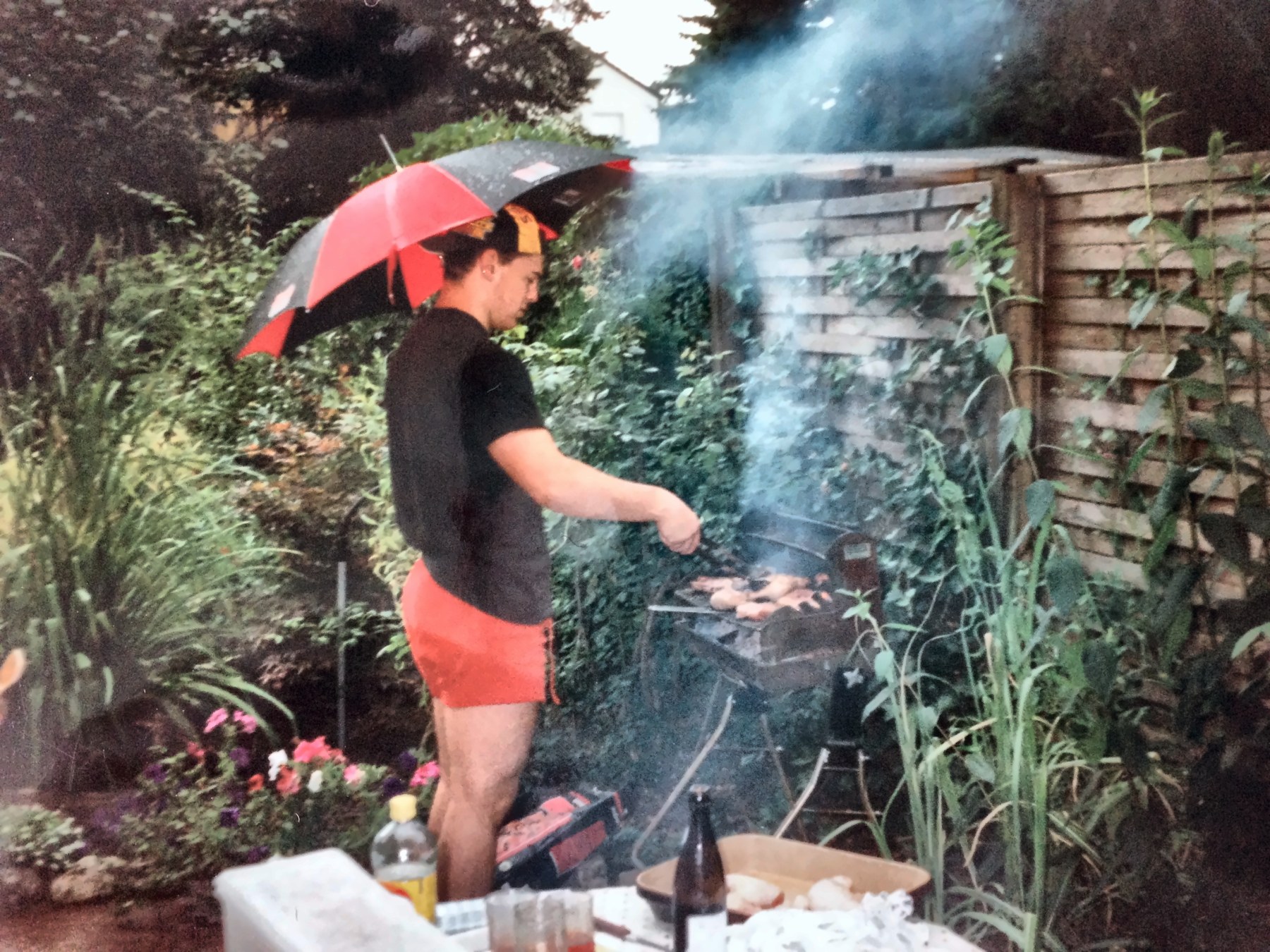 Tom grillt in seiner Jugend unter einem Regenschirm an einem Holzkohlegrill im Garten