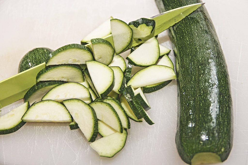 Zucchini wird auf einem Brett mit dem Messer in Scheiben geschnitten, daneben eine ganze Zucchini