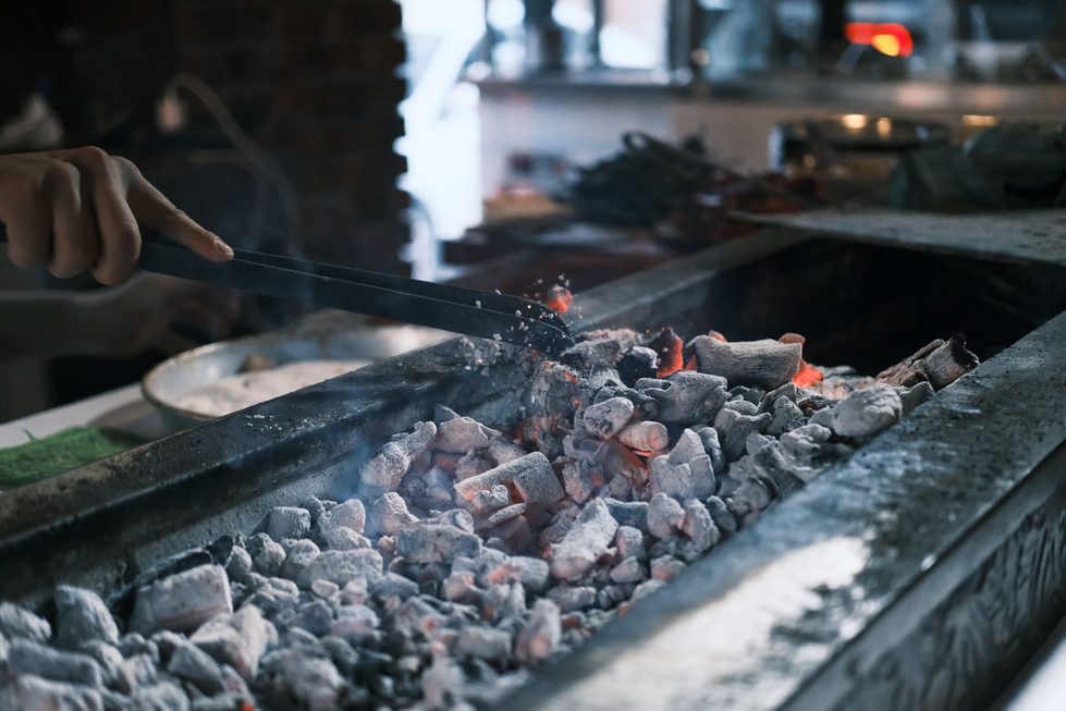 Eine Hand verteilt mit einer Grillzange durchgeglühte Holzkohle mit grauer Ascheschicht im offenen Grill, keine Lebensmittel sichtbar.