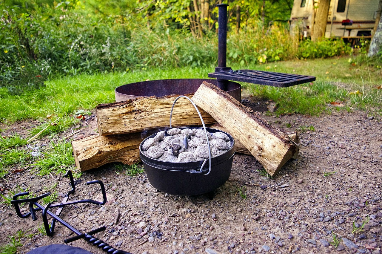 Gusseiserner Dutch Oven mit durchgeglühten Briketts auf dem Deckel, daneben Holzscheite und ein Deckelheber