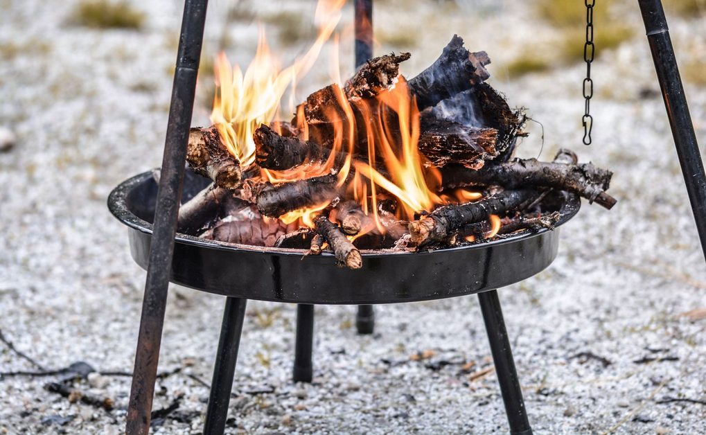 Eine schwarze Feuerschale steht auf mineralischem Untergrund im Garten, darüber ist ein stabiles Dreibein aufgebaut, in der Schale lodert ein Feuer.