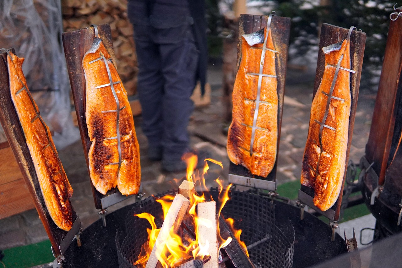 Lachsseiten auf senkrecht stehenden Holzbrettern rund um einen Feuerkorb mit brennenden Holzscheiten