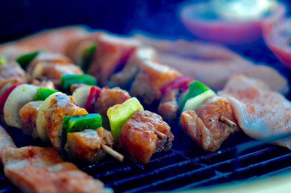 Bunte Gemüse- und Hähnchenspieße, Fischfilets und mageres Grillfleisch liegen nebeneinander auf dem Grillrost in sommerlicher Atmosphäre.