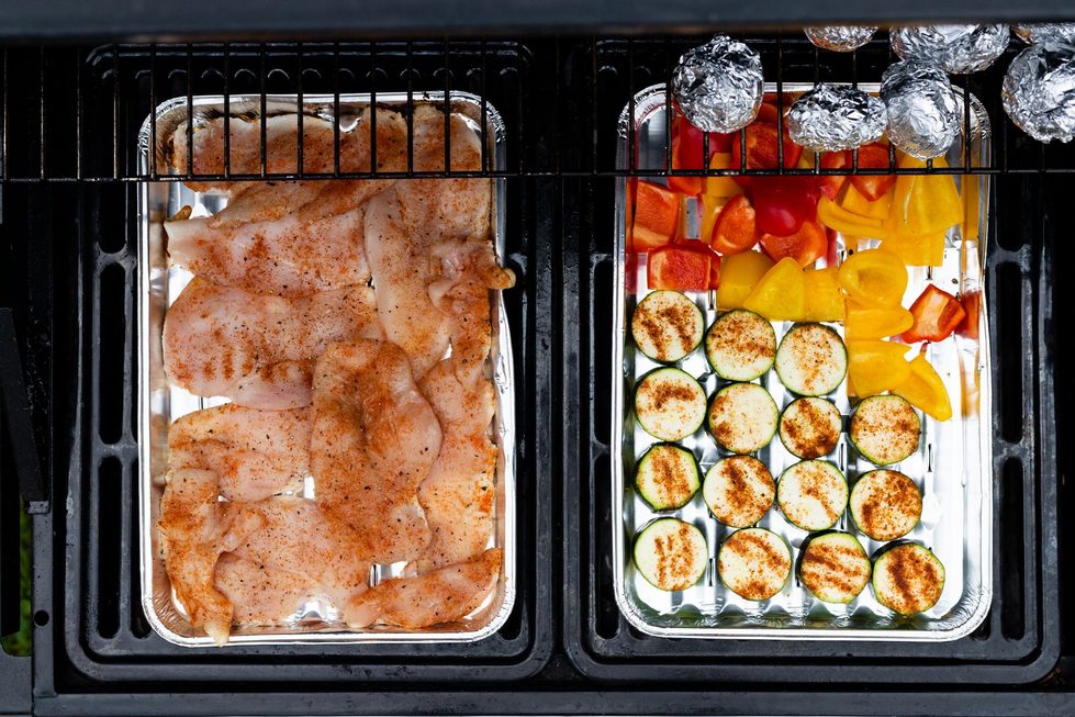 Blick von oben auf zwei Grillzonen nebeneinander, eine mit gewürztem Fleisch in einer Schale, die andere mit Gemüse und Kartoffeln beim Grillen indirekt.