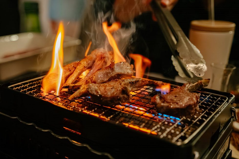 Lammkoteletts liegen auf einem Grill über offenen Flammen, eine Grillzange hält ein Stück, Rauch zieht auf, Hintergrund unscharf.
