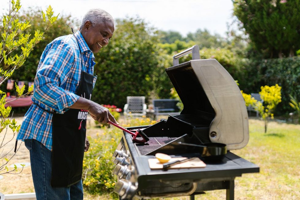 Ein Mann grillt entspannt an einem Gasgrill im grünen Garten, im Hintergrund stehen Gartenmöbel und Pflanzen bei warmem Sommerwetter.