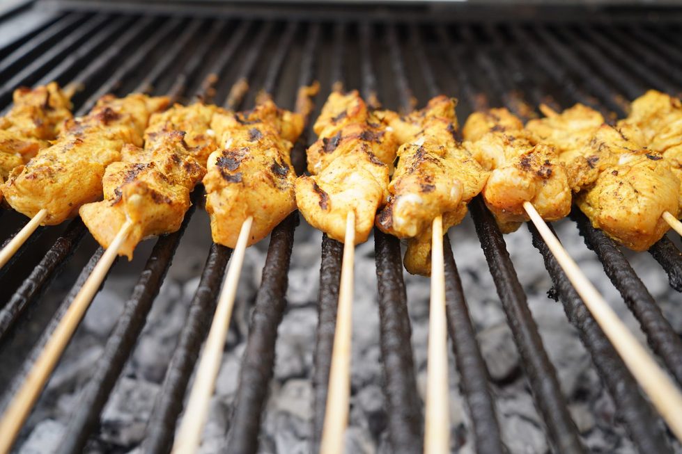 Nahaufnahme von mehreren goldbraun gegrillten Hähnchenspießen auf Holzspießen, die über glühender Kohle auf einem Grillrost liegen.