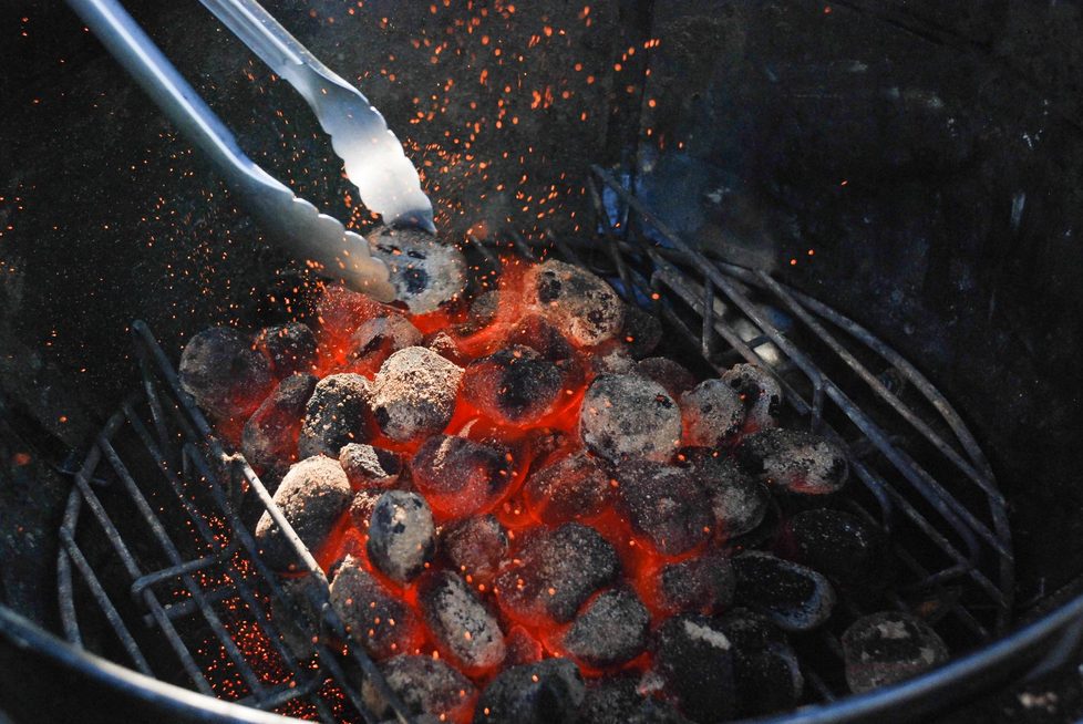 Nahaufnahme von glühender Holzkohle auf einem Kohlerost im runden Grill, mit Grillzange und Funkenflug, im Hintergrund Grillwand.