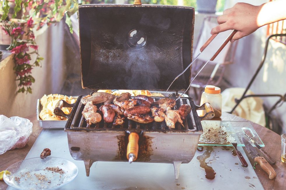Outdoor-Grill mit verschiedenen Stücken Geflügel auf dem Rost, Grillzange und sauberer Arbeitsfläche im sonnigen Garten.