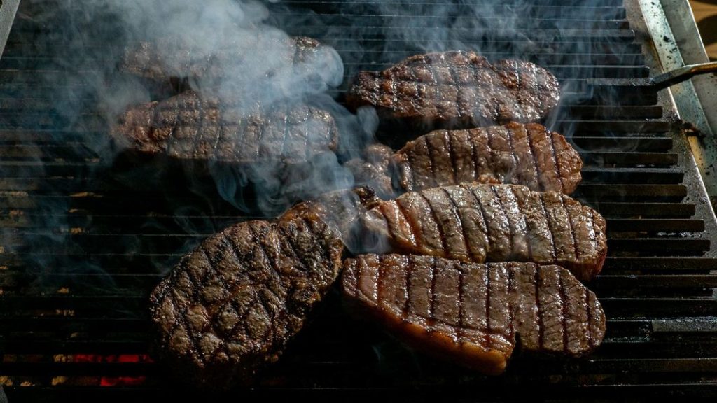 Mehrere dicke Rindersteaks liegen auf einem heißen Grillrost, darüber steigt Rauch auf.
