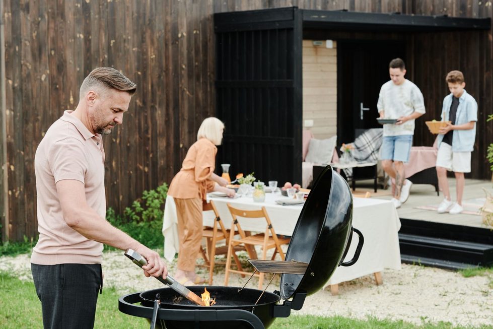 Ein Erwachsener überwacht einen offenen Grill im Garten, während Kinder weit entfernt an einem festlich gedeckten Tisch spielen.