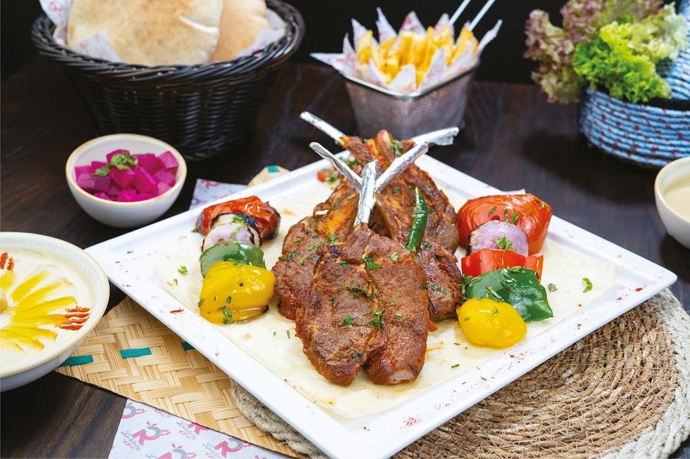Platte mit gegrillten Lammkoteletts, buntem Grillgemüse, Fladenbrot, Salat und Joghurt, dekorativ angerichtet auf einem Tisch.