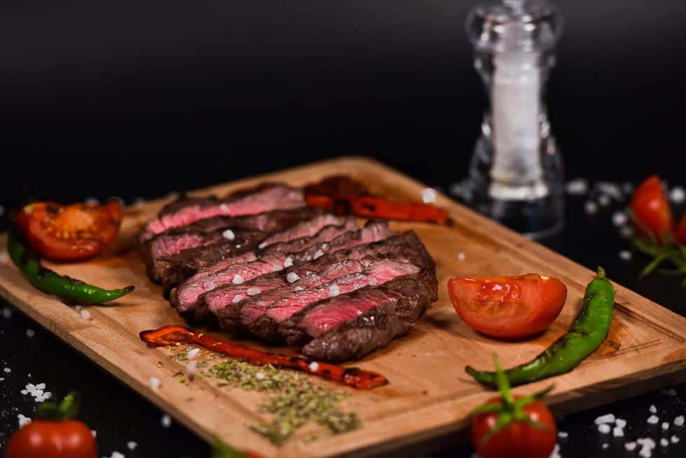 Aufgeschnittenes gegrilltes Rindersteak mit kräftig rosa Kern auf einem Holzbrett, daneben Tomaten und grüne Chili.