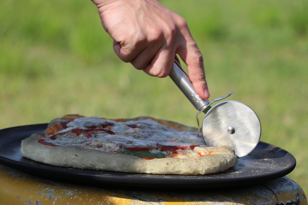 Selbstgemachte Pizza auf einer runden dunklen Platte wird im Freien mit dem Pizzaschneider geschnitten