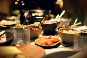 Gedeckter Raclette-Tisch mit Schälchen voller Zutaten, rohem Lachs auf einem Teller und dem Raclettegerät im Hintergrund