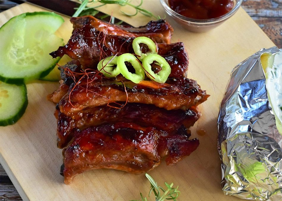 Portionierte, glasierte Spare Ribs liegen auf einem Holzbrett mit frischen Gurkenscheiben, grüner Chili, Salat und Backkartoffel.