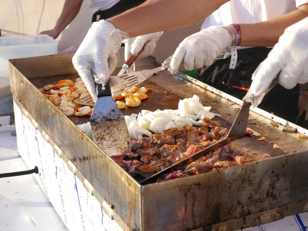 Zwei Spachtel wenden Fleischstücke, Zwiebeln und Garnelen auf einer großen heißen Grillplatte