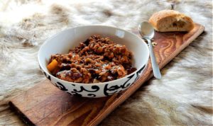 Eine Schale Chili con Carne mit Hackfleisch und Bohnen steht mit Löffel und einem Stück Brot auf einem Holzbrett über einem Fell.