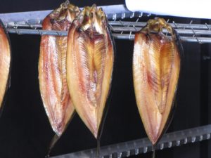 Drei aufgeklappte, goldbraun geräucherte Fische hängen an Haken nebeneinander in einem Räucherofen.
